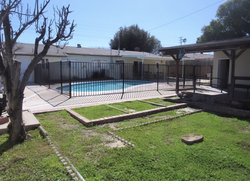 A backyard featuring a fenced swimming pool, green lawn, and patio area.