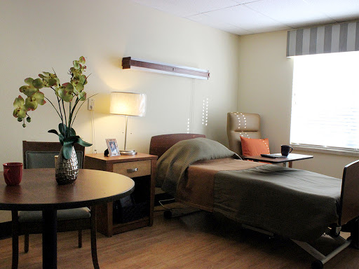 A serene and well-decorated hospital patient room, featuring a bed, bedside table with a lamp, and a small dining area.