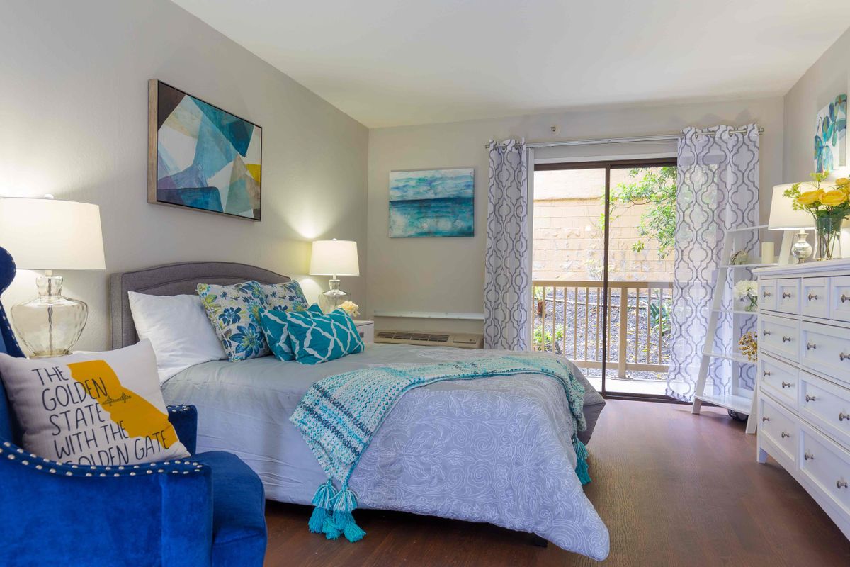 A cozy bedroom featuring a neatly made bed with colorful pillows and throws, artwork on the walls, and natural light streaming in through sliding glass doors leading to a balcony.