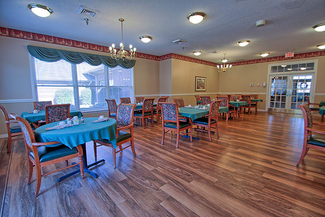 A spacious dining room with neatly arranged tables and chairs, featuring elegant chandeliers and large windows with curtains.