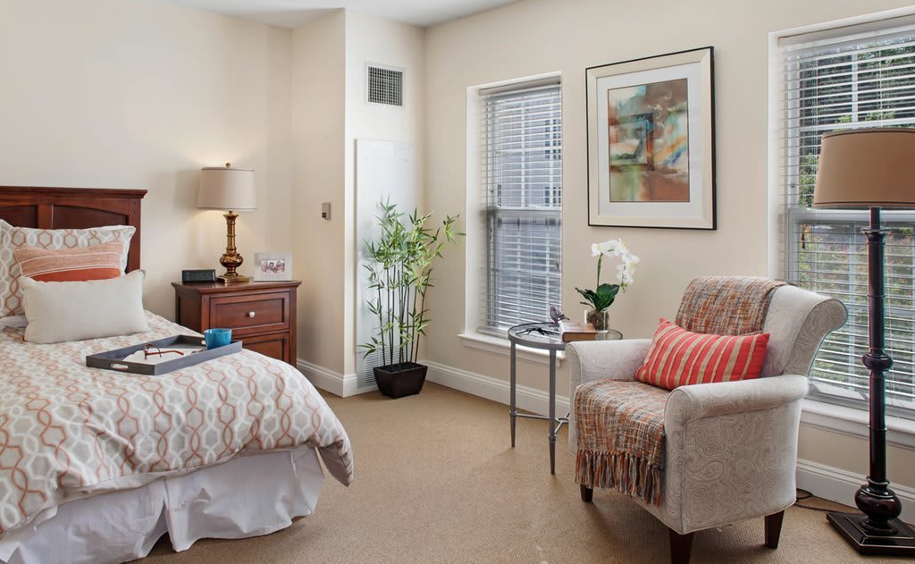 A cozy bedroom with elegant decor, featuring a bed, armchair, side table, and potted plants.