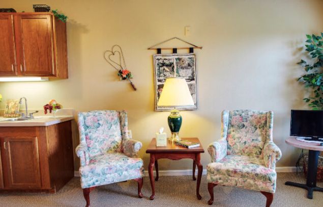 A cozy living room setup with two floral-patterned chairs, a wooden side table, and a table lamp, accompanied by wall decor.