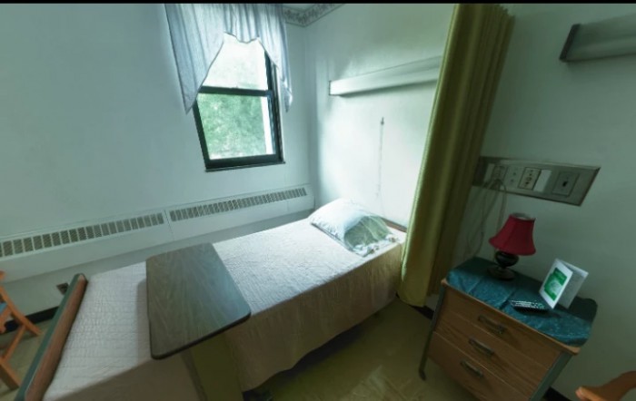 A small, simple room with a single bed next to a window, accompanied by a bedside table and dresser.