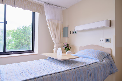 A serene and well-lit hospital patient room featuring a bed with a light blue blanket, a small table, and a window adorned with curtains, complemented by a vase of fresh flowers.