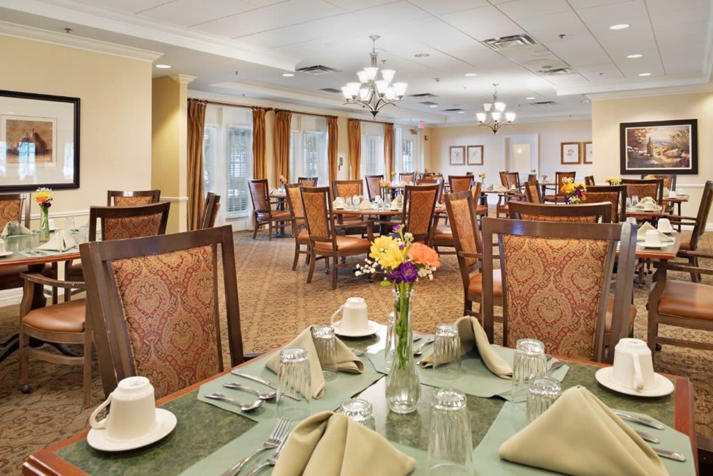 A spacious dining room with elegantly set tables and comfortable chairs, featuring floral centerpieces.