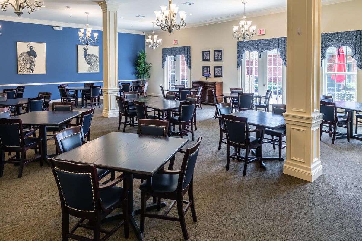 A spacious dining area with multiple tables and chairs, featuring elegant chandeliers and a carpeted floor, complemented by blue accent walls and large windows.