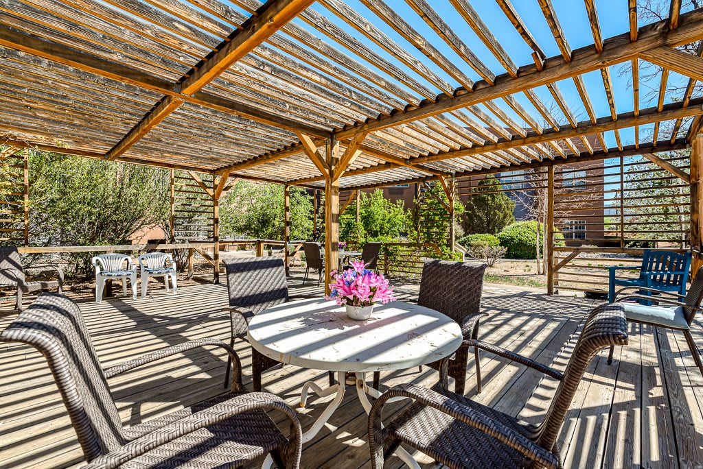 A cozy outdoor patio with a wooden pergola, providing shade over tables and chairs on a sunny day.
