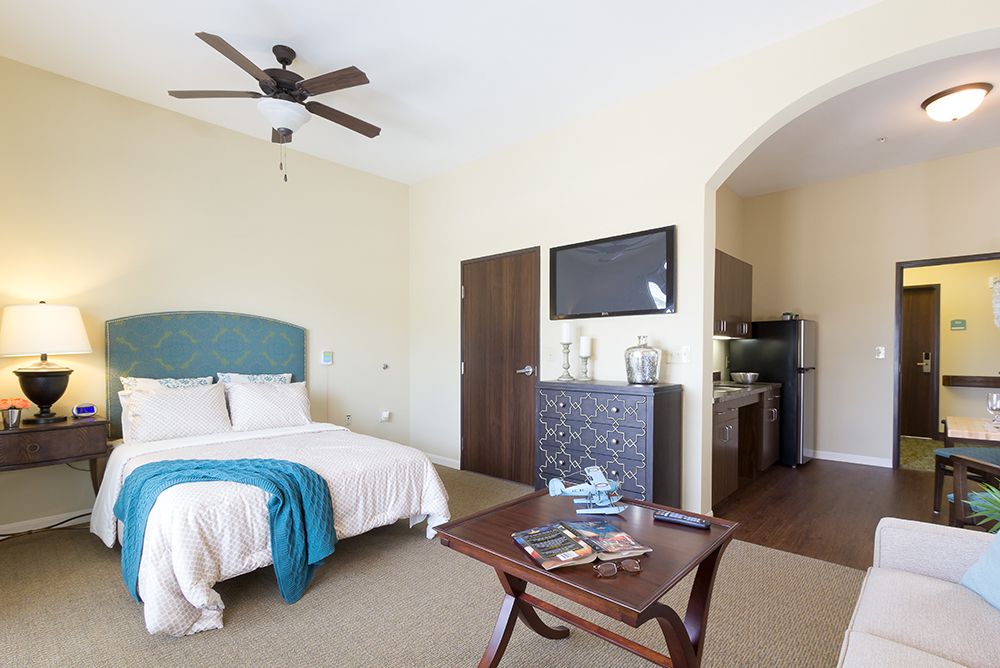 A modern and spacious hotel room featuring a cozy bed, ceiling fan, television, and a small kitchenette.