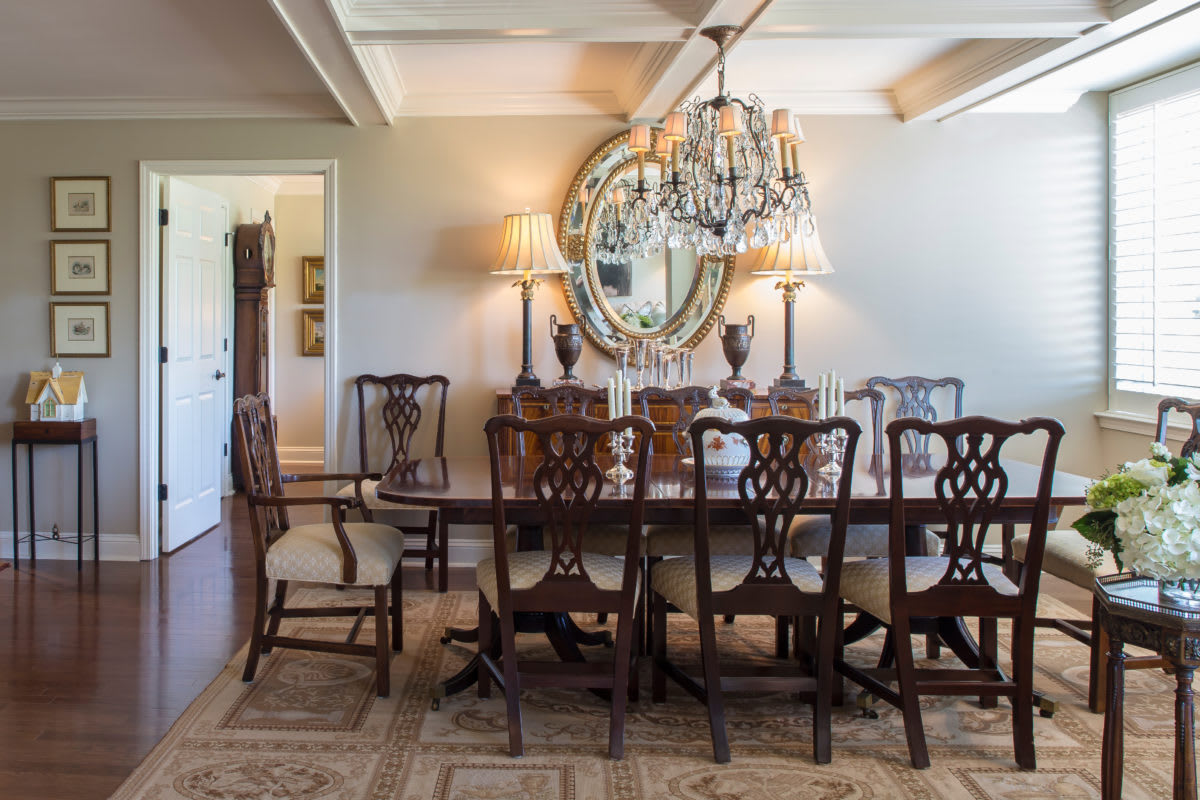 A traditional dining room featuring a crystal chandelier, a long wooden table with matching chairs, and an ornate mirror on the wall.