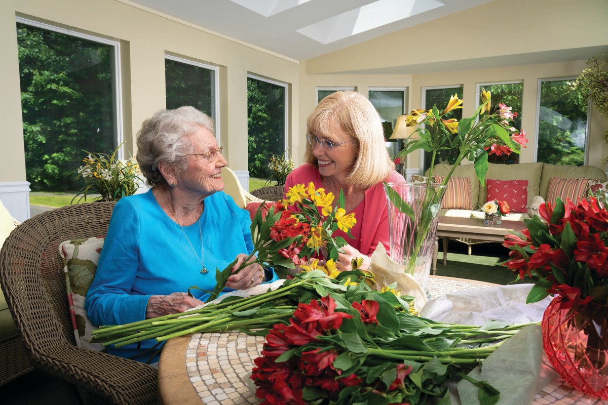 An elderly woman and a younger woman smile as they arrange flowers together indoors.