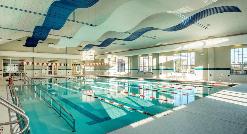 A spacious indoor swimming pool with lane dividers and a wave-themed ceiling design, illuminated by abundant natural light.
