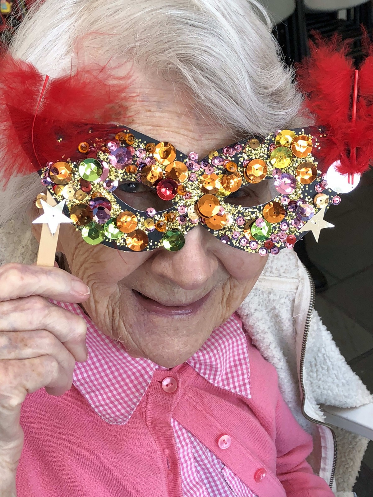 An elderly person wearing a colorful, glittery mask adorned with feathers.