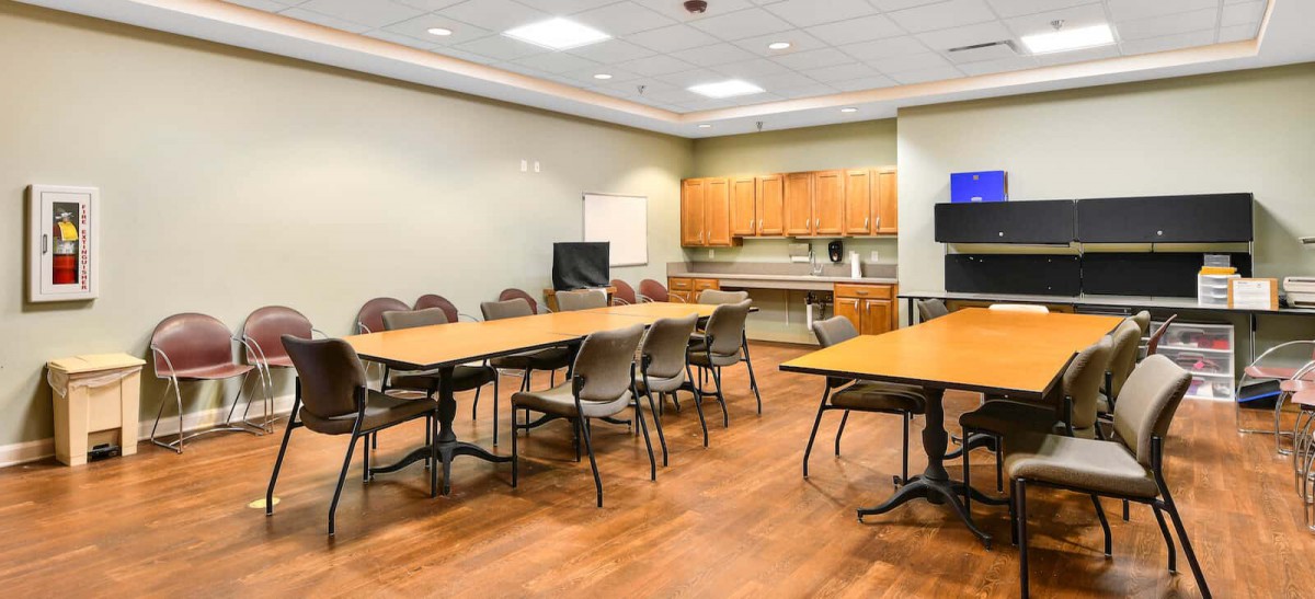 A spacious conference room with tables and chairs arranged for a meeting, featuring a TV screen and whiteboard.
