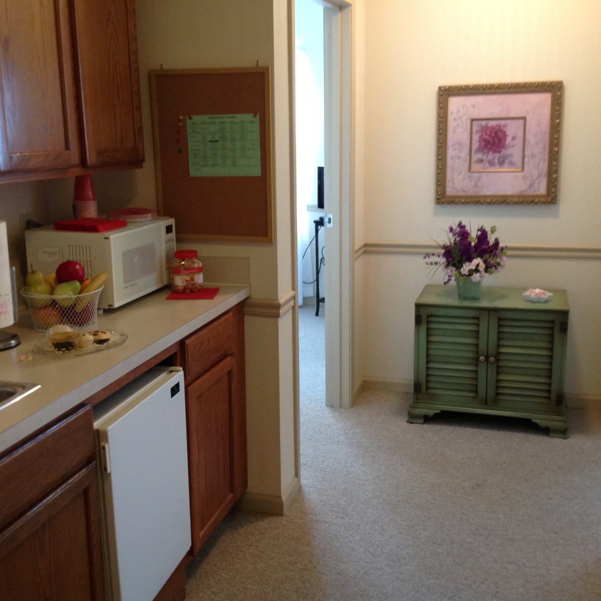A cozy kitchen area with wooden cabinets, a microwave, and decorative elements like flowers and artwork.