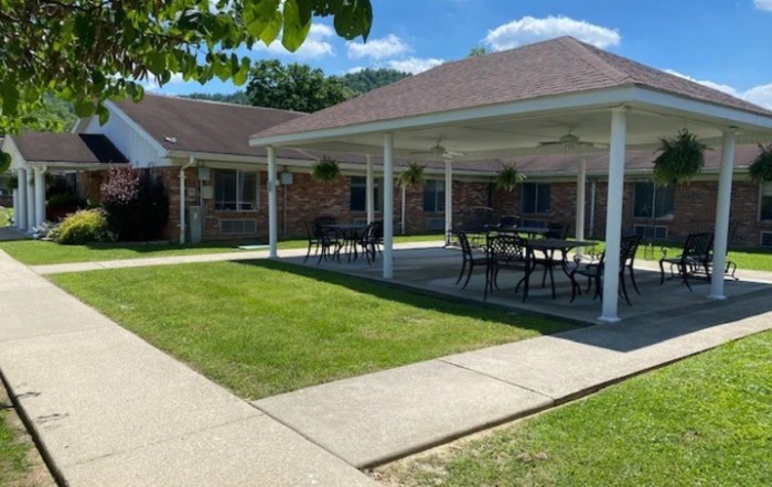 A covered patio area with tables and chairs next to a building, surrounded by a green lawn.
