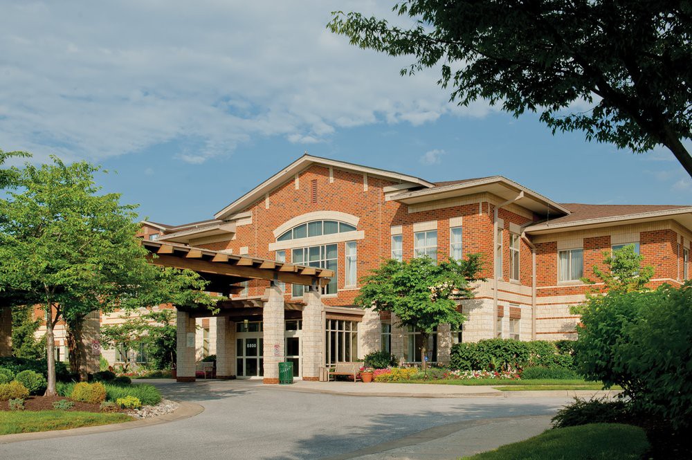 A two-story brick building with a prominent entrance, surrounded by trees and set against a clear blue sky.