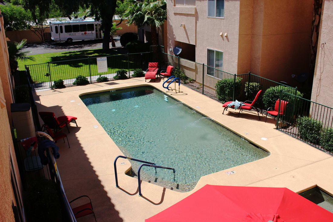 An outdoor swimming pool at an apartment complex on a sunny day, surrounded by lounge chairs and a safety fence.