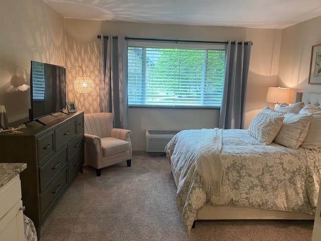 A cozy bedroom with a large bed, patterned bedding, and elegant decor featuring a chair, dresser, and ample natural light from the window.