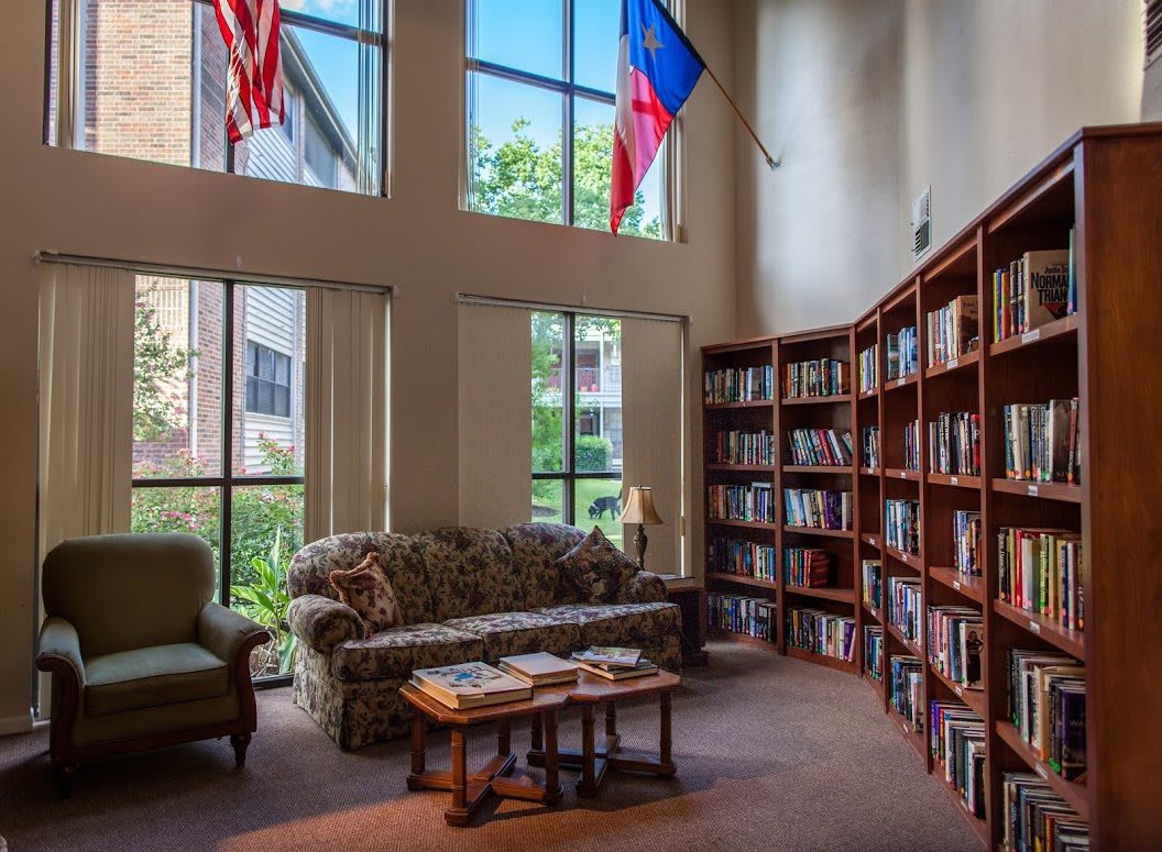 A cozy library corner with a sofa, armchair, and bookshelves under tall windows and flags.