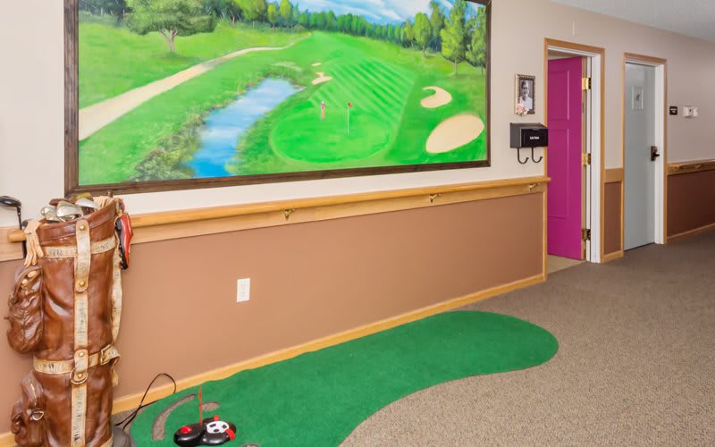 An indoor hallway features a putting green and a vibrant golf course mural on the wall.