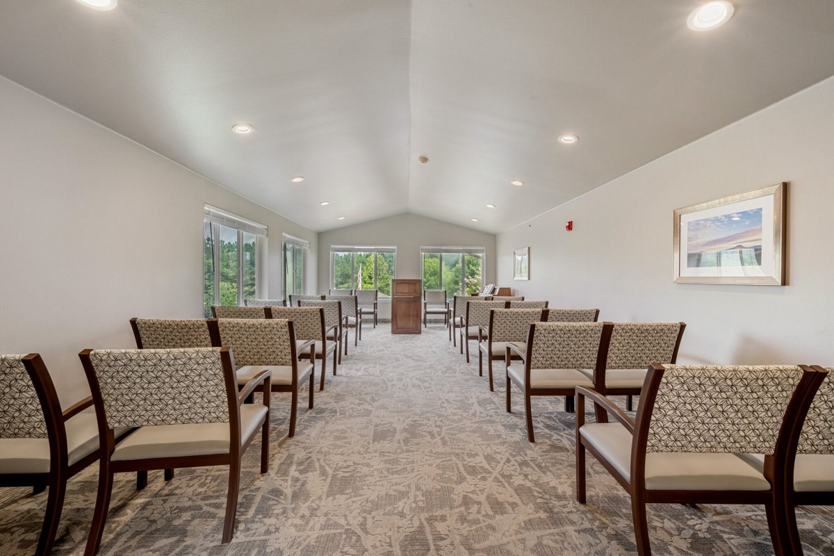 A bright and spacious room with arranged chairs and a podium, featuring large windows and soft lighting.