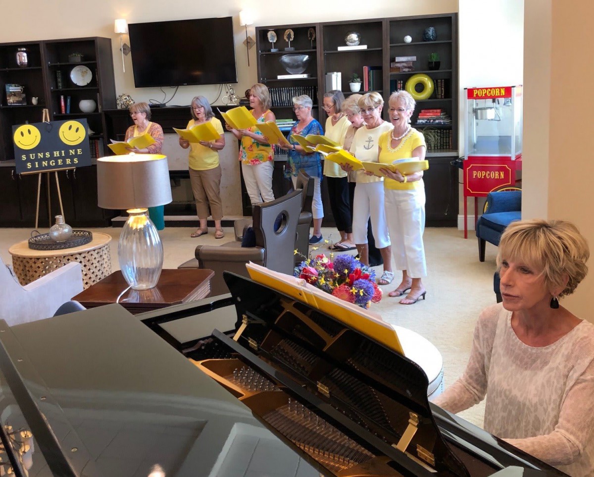 A group of women singing in a choir while one plays the piano indoors.