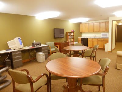 A spacious office break room with tables, chairs, a computer setup, and a kitchenette.
