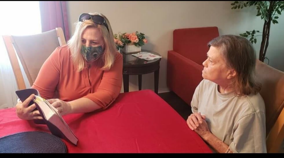 Two individuals sitting at a table, engaging in conversation. One is wearing a mask and holding something.