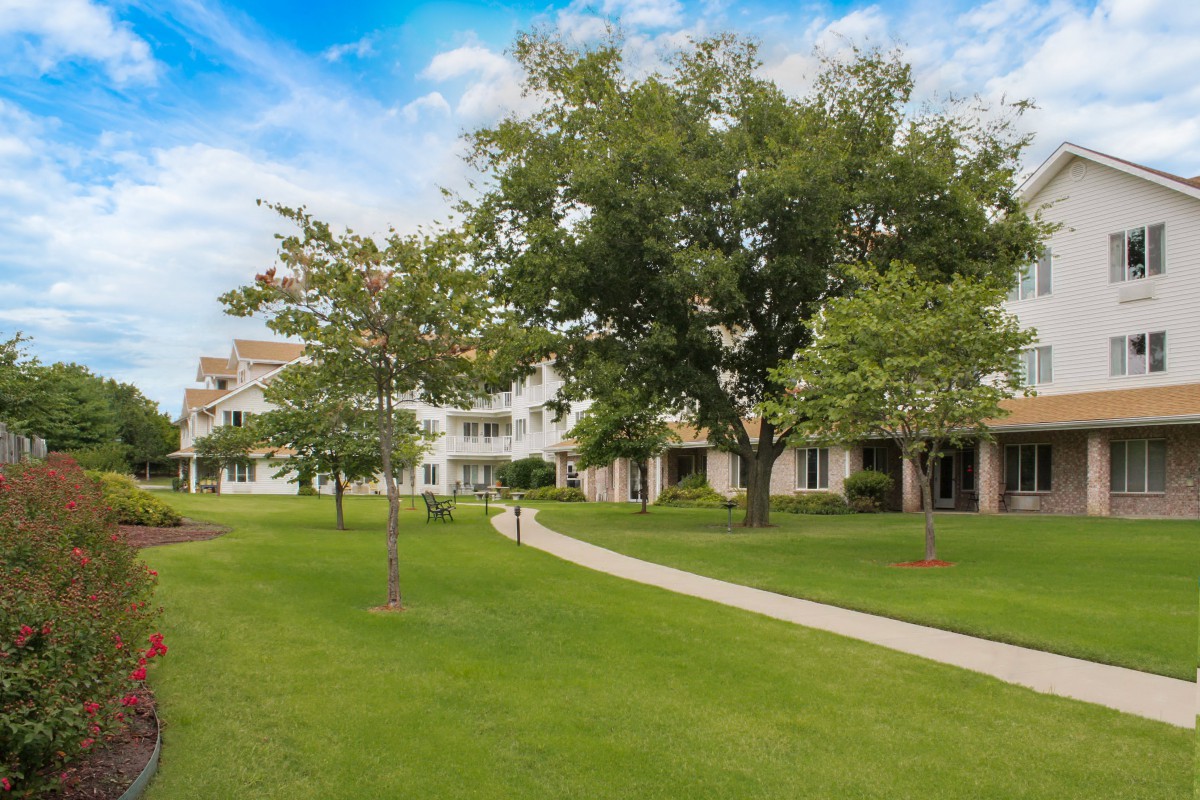 A scenic view of a grassy area with a walkway, surrounded by trees and residential buildings.