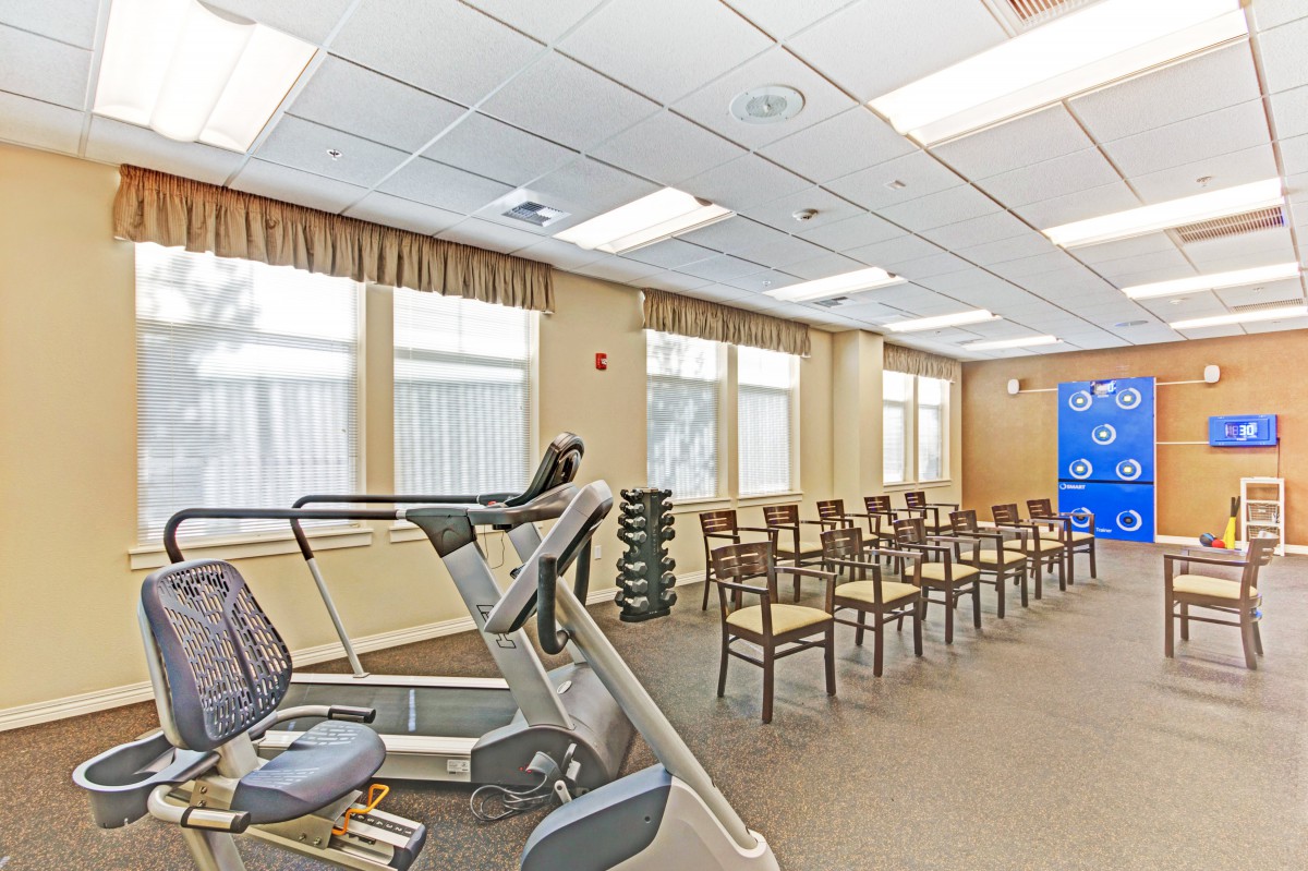 A multipurpose room with gym equipment and rows of chairs, illuminated by natural and ceiling lights.