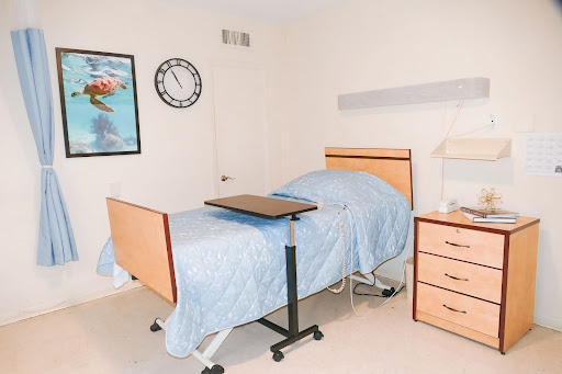A clean and organized hospital room featuring a patient bed with a blue blanket, a bedside table, and a wall clock.