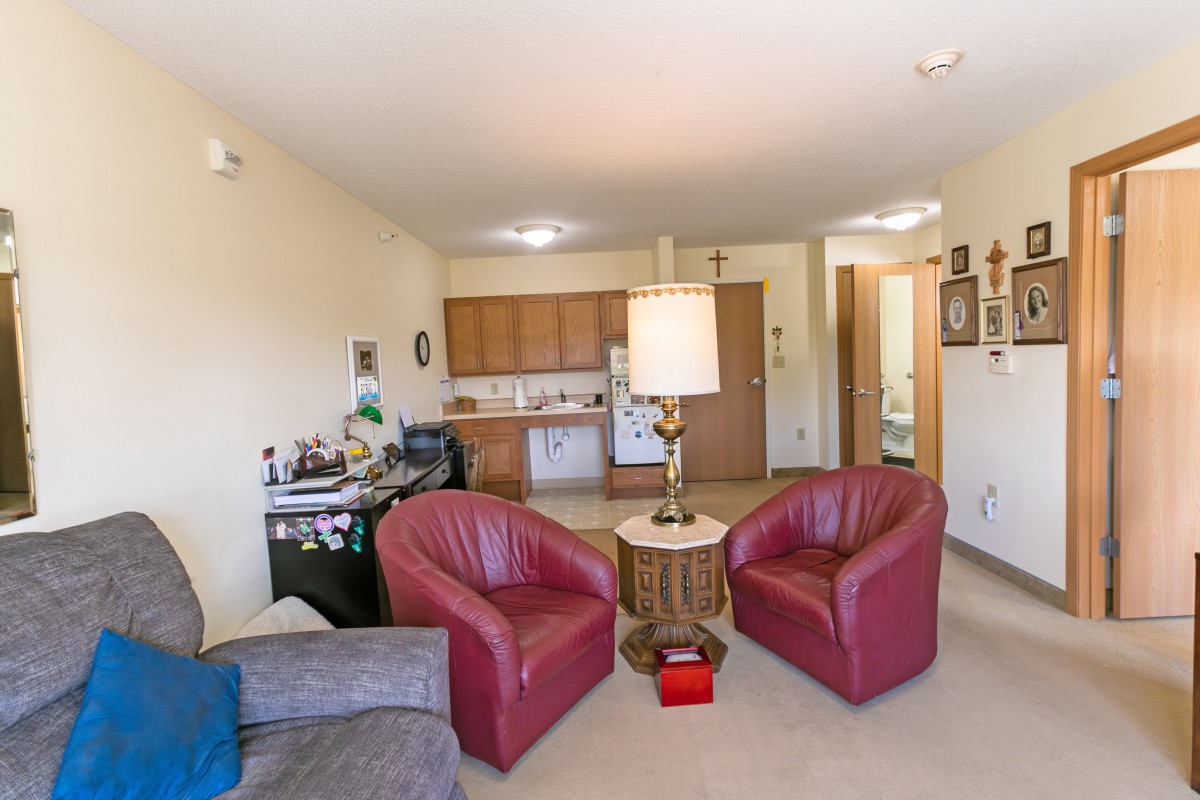 A cozy living room with red armchairs, a central lamp on a round table, and an adjacent kitchen area.
