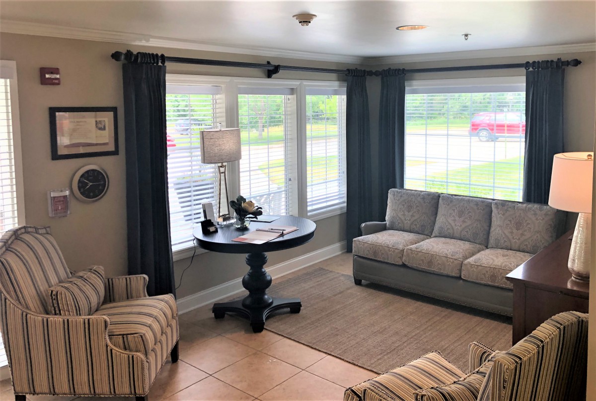 A cozy living room with large windows, featuring a sofa, armchair, and a round table with a lamp.