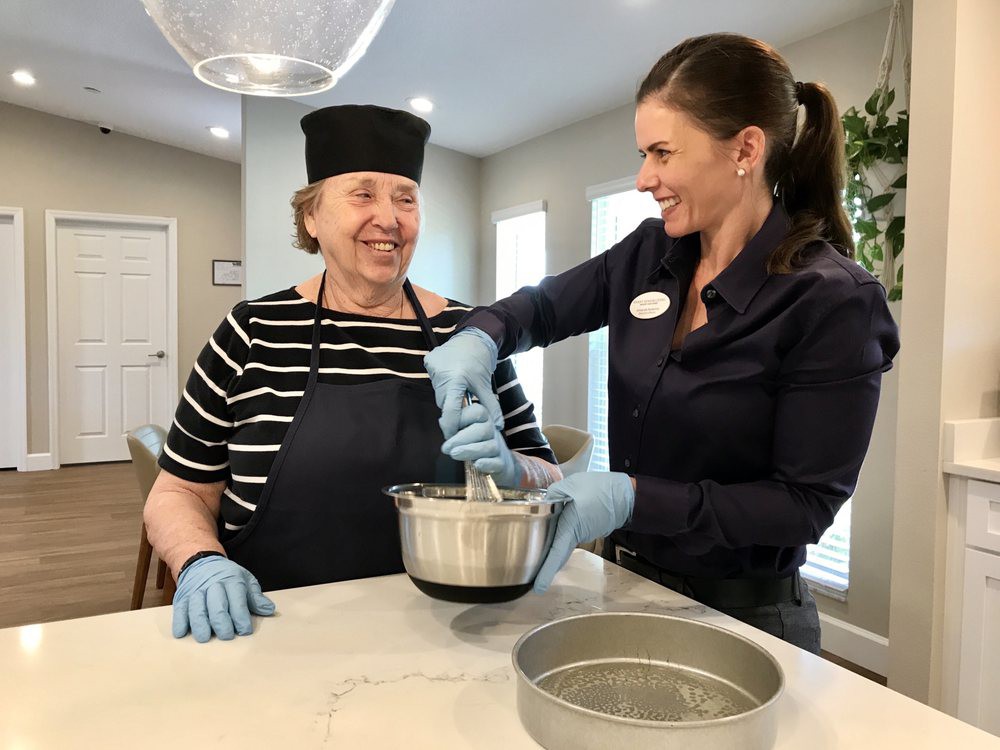 An elderly woman in a chef hat and apron smiles while a younger woman helps her mix ingredients in a kitchen.