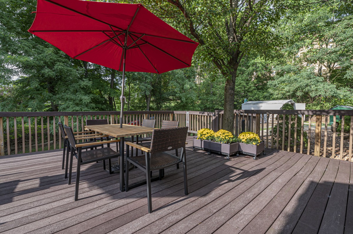 A cozy patio deck with a table and chairs under a red umbrella surrounded by greenery.