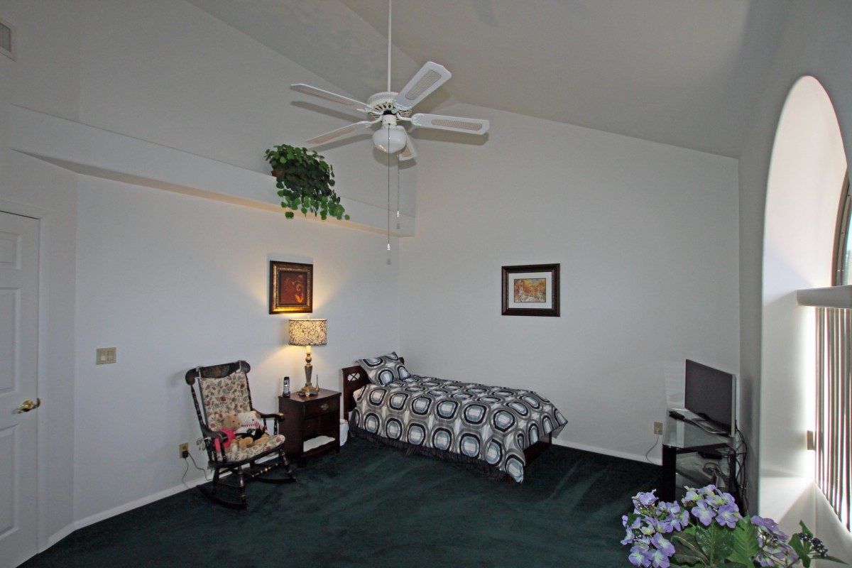 A cozy bedroom with a single bed, featuring a lamp, chair, and ceiling fan.