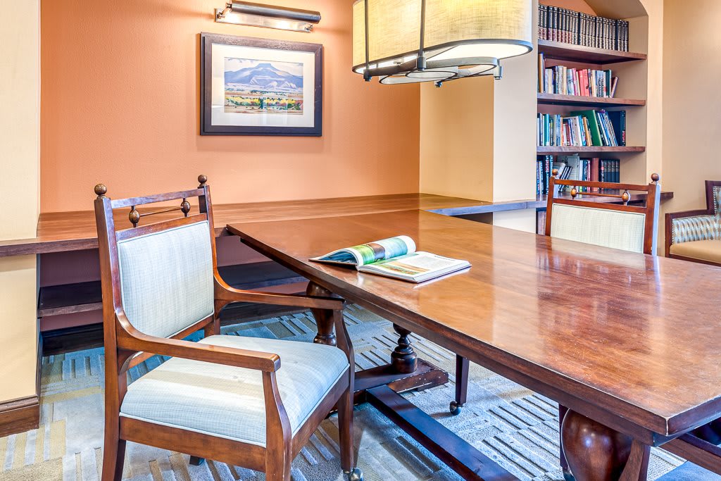 A cozy library reading room featuring a wooden table, chairs, bookshelves, and artwork on the wall.