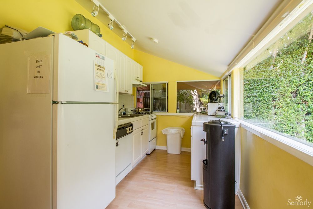 A bright kitchen with yellow walls and wooden flooring, featuring a refrigerator, cabinets, and ample natural light from large windows.