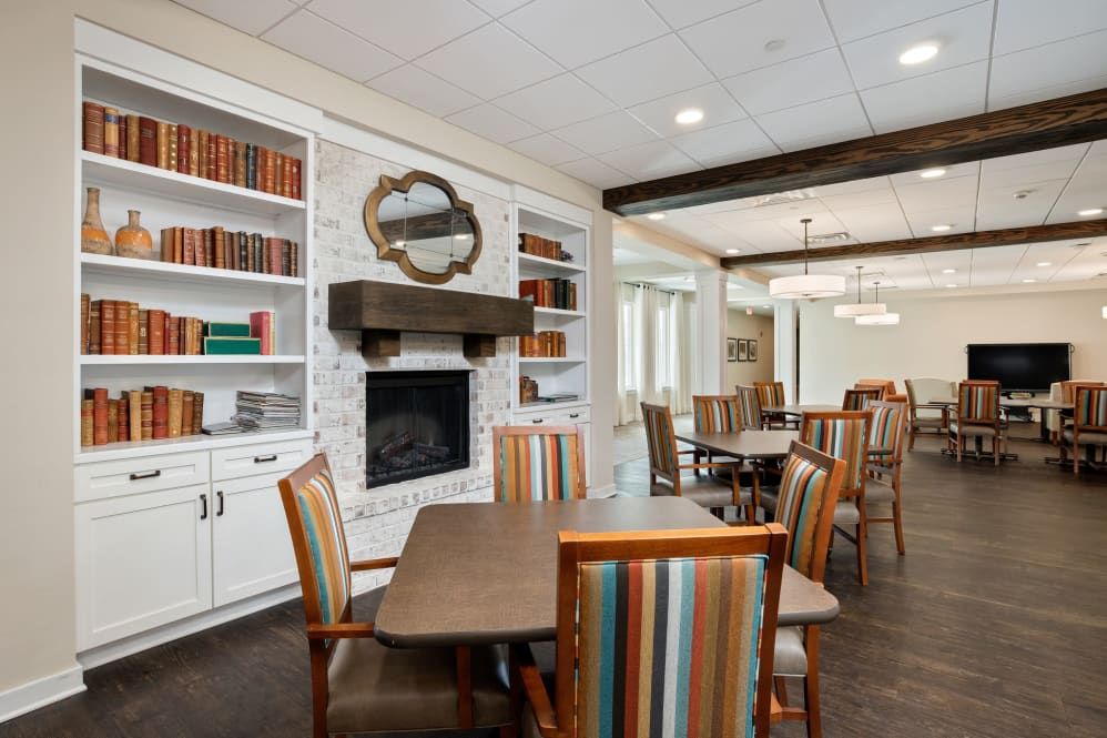 A cozy library and dining area with bookshelves, a fireplace, and colorful chairs around wooden tables.
