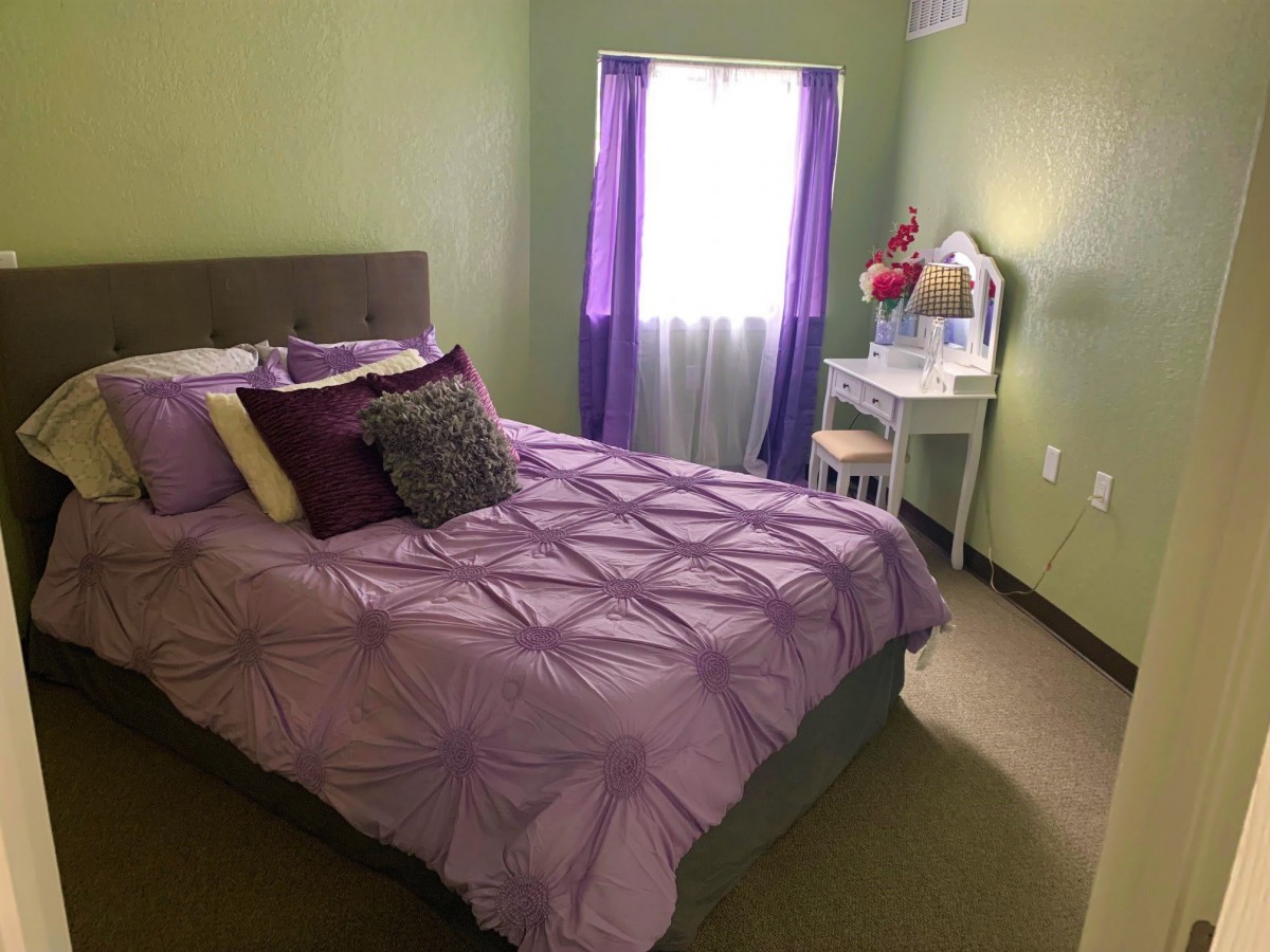 A cozy bedroom with purple bedding and green walls, featuring a window with light streaming in and a vanity table by the side.