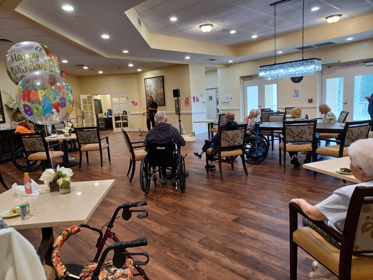 Residents gather in a common area of a nursing home, with some in wheelchairs and others seated around tables.
