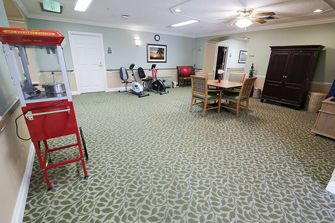 A spacious living room with carpeted flooring, featuring a television, chairs, and a table. People are seated and engaged in activities.