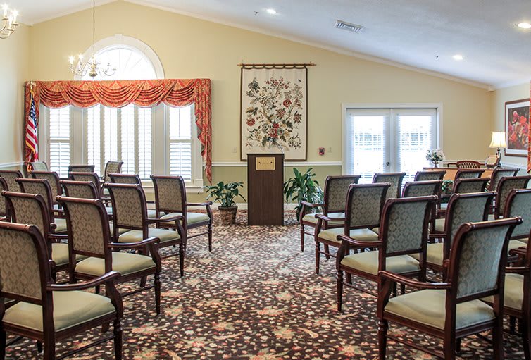 An interior view of a room set up with rows of chairs facing a podium, featuring decorative plants and a patterned carpet.