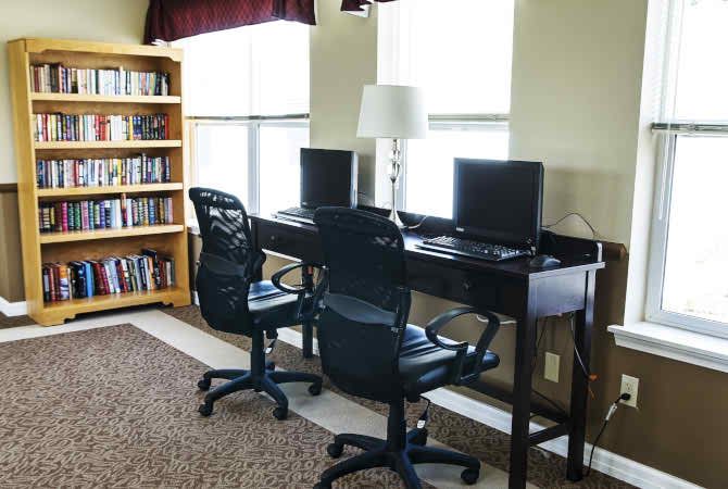 A small library area featuring a bookshelf, two computers on a desk, and comfortable chairs by the windows.