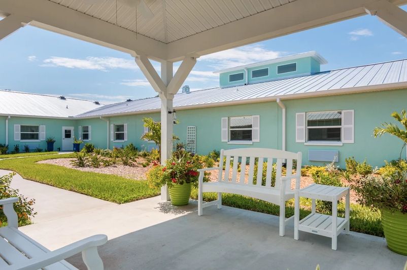 A serene outdoor area featuring a white bench under a covered porch, with pastel-colored buildings and a garden in the background.