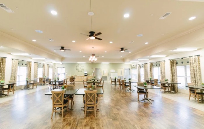 A spacious dining hall with neatly arranged tables and chairs, featuring wooden flooring and ceiling fans.