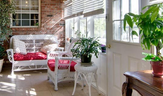 A cozy sunroom featuring wicker furniture, bright red cushions, and a potted plant basking in natural light.