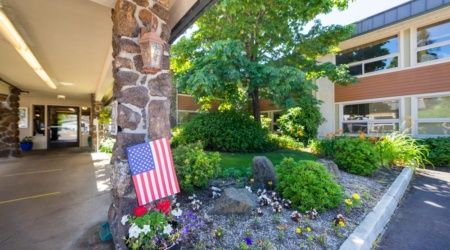 A garden scene featuring an American flag placed near a stone pillar surrounded by lush greenery and flowers.