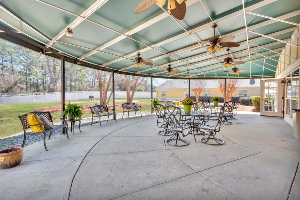 A spacious covered patio with tables and chairs, featuring ceiling fans and potted plants, overlooking a backyard.