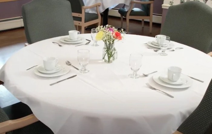 A neatly set round dining table with white tablecloth and a small floral centerpiece.
